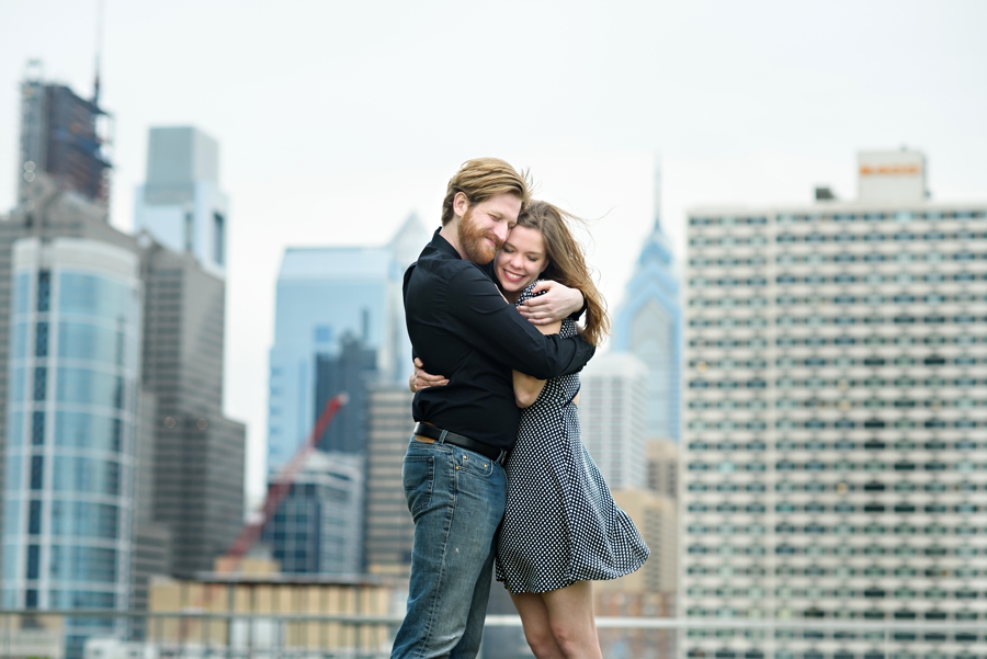 An engaged couple wrap each other up in their arms with the philadelphia skyline behind them during their engagement session.